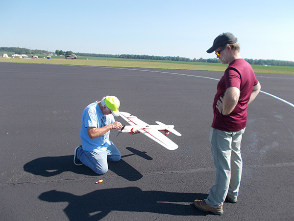 Man kneeling adjusts model airplane on runway, another stands watching. Bright sunny day.
