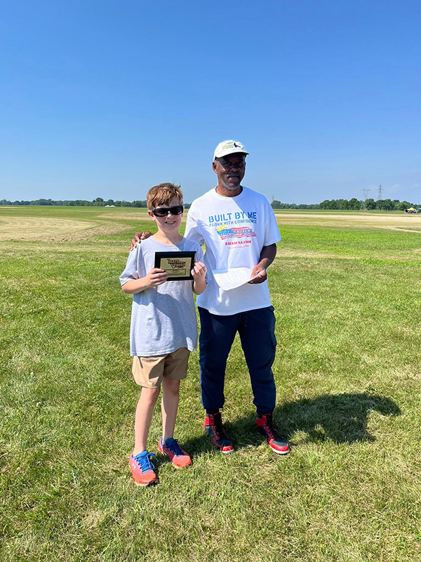 A boy holding a plaque stands next to an adult on a grassy field, both smiling.