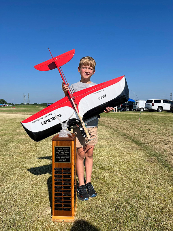 Young boy holding a red-and-white model airplane next to a trophy on a sunny day.