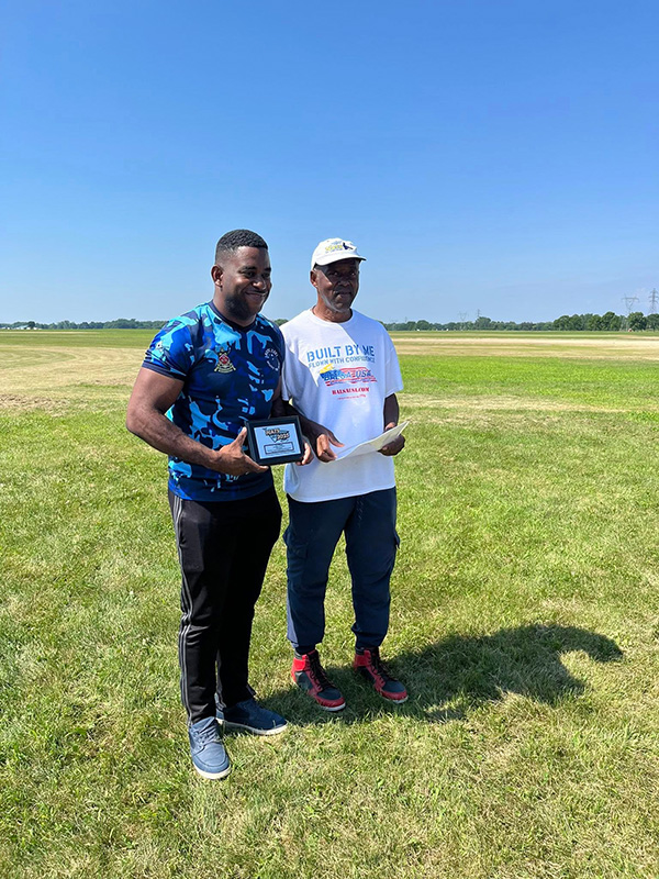 Two men standing on grass, one holding a plaque, under a clear blue sky.