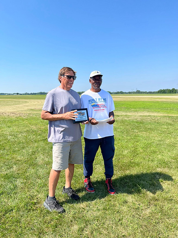 Two men standing on grass under a clear blue sky, one holding a plaque.