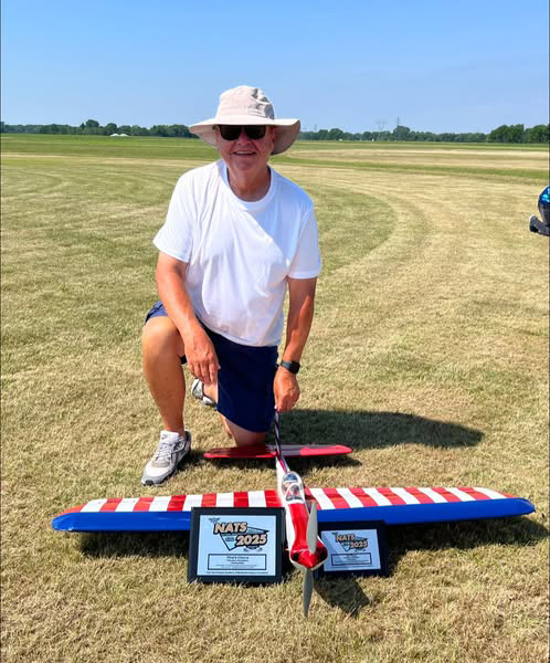 Man kneeling on grass beside a red, white, and blue model plane with awards.