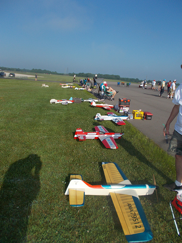 Model airplanes lined up on grass alongside a runway under a clear blue sky.