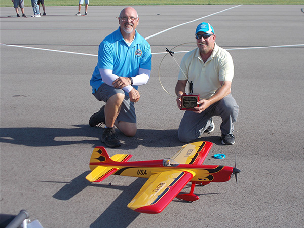 Two men kneeling on pavement with a large yellow and red model airplane.