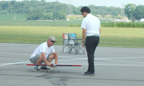 Man crouching by a small model airplane on a runway, another man standing beside him.