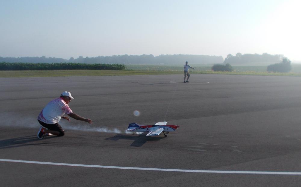 Two people on an airstrip with a model plane, surrounded by open fields.