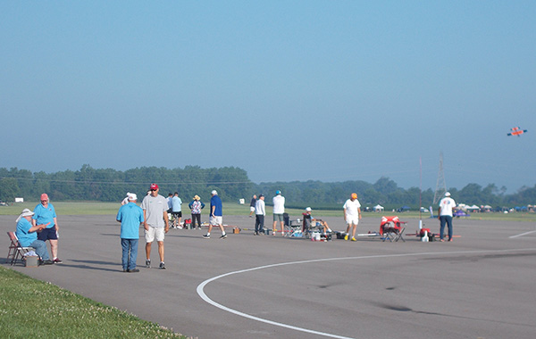 People gathered on an airfield, with a small plane flying in the background.