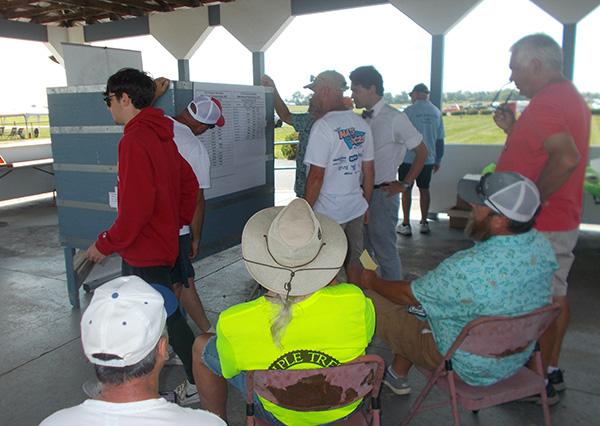 People discussing by a bulletin board in a shaded outdoor area.