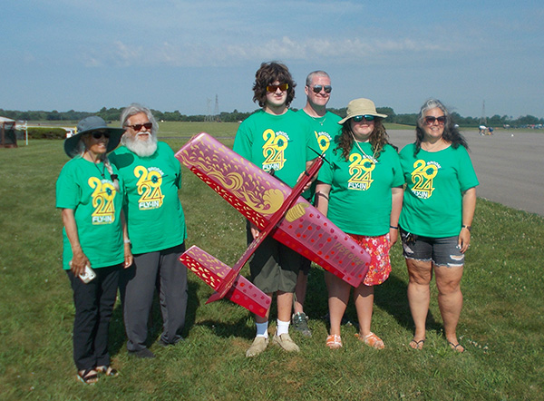 Group with red model plane, wearing matching green shirts, outdoors on grass.
