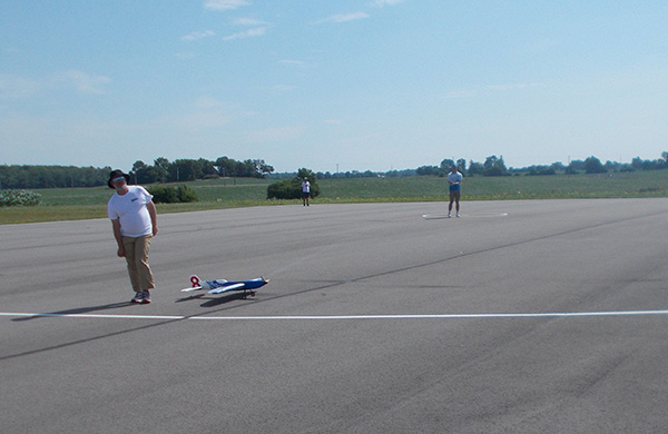 Remote-controlled airplane on a tarmac, people standing nearby under blue sky.