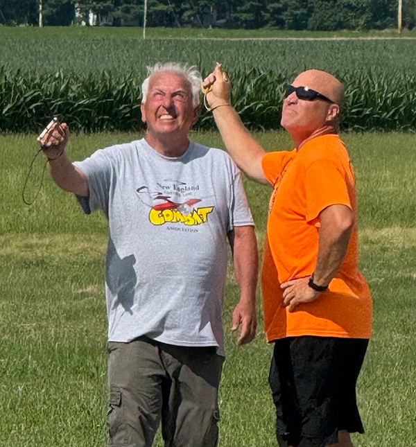 Model airplane taking off on paved runway, two people in background.
