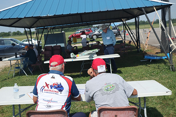 People sitting under a canopy at an outdoor event with model aircraft.