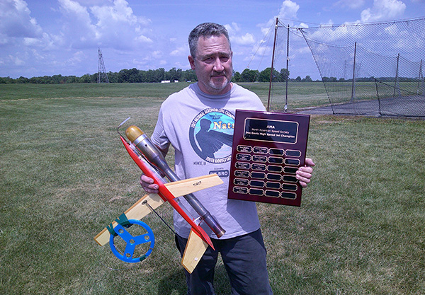 Man holding a rocket and plaque on grassy field.