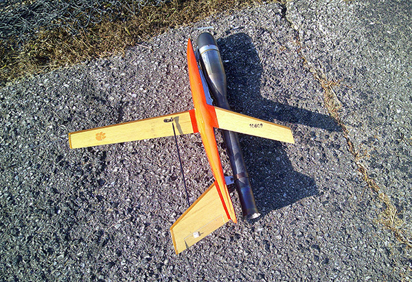 Model rocket glider on asphalt, with orange and wooden wings.