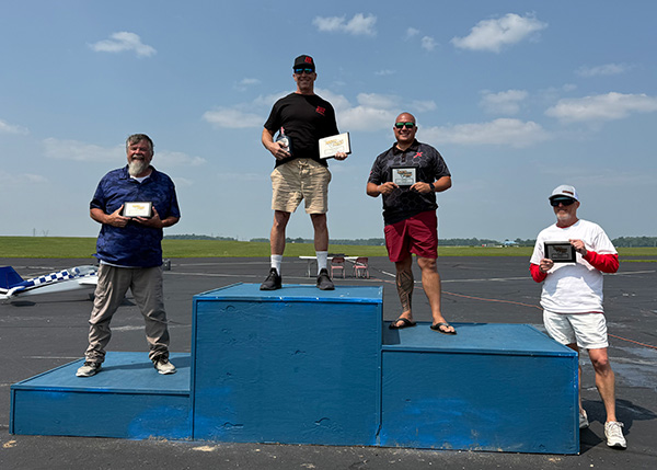 Five men standing on winners&apos; podium outdoors under a clear sky.
