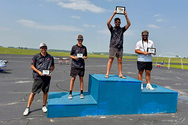 Four men holding plaques stand on a winners&apos; podium outdoors, under a clear blue sky.