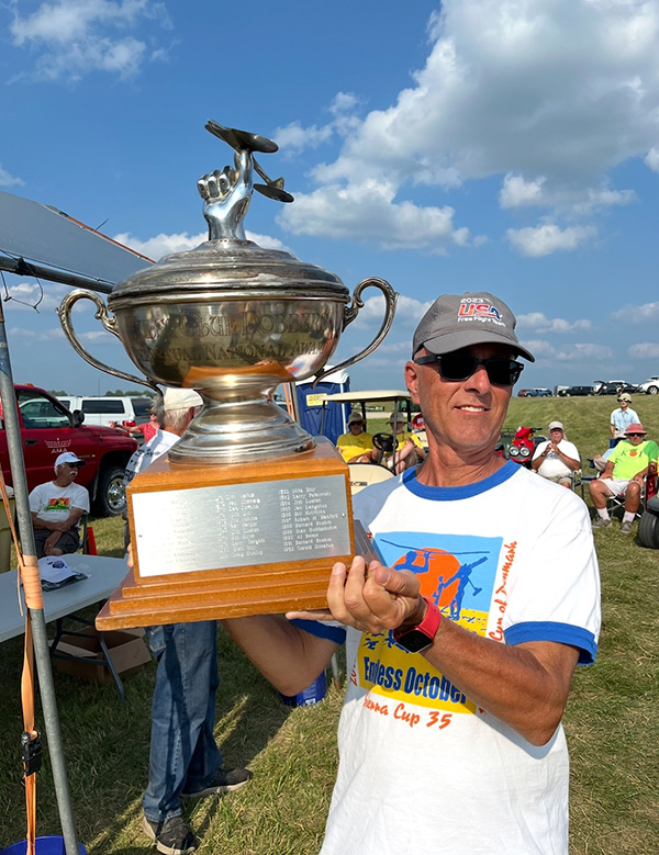 Man holding large silver trophy outdoors, wearing sunglasses and a cap.