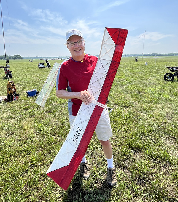 Man smiling, holding a large red-and-white model airplane in a grassy field.