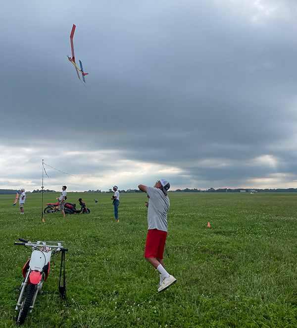 Man in shorts flying a model airplane in a grassy field, with cloudy sky.