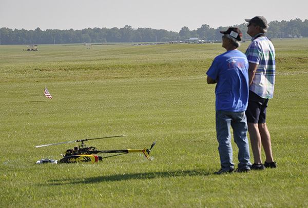 Remote-controlled helicopter on grass, two men standing nearby watching.