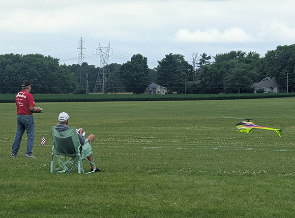Two men watching a remote-controlled helicopter fly over a grassy field.