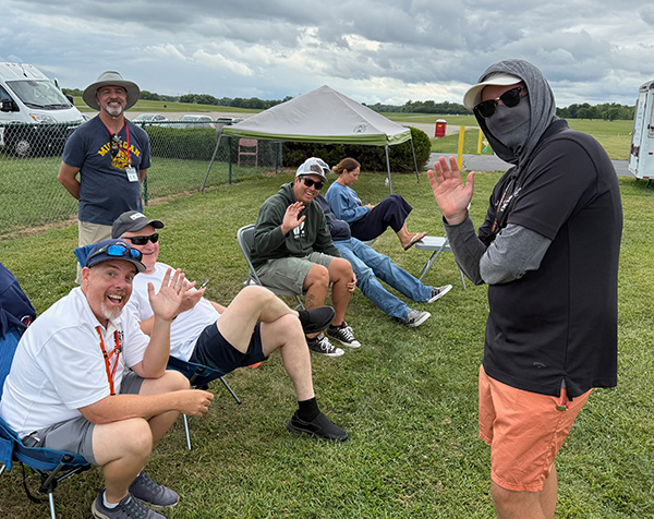 Group of people smiling and waving on a grassy field, with tents in the background.