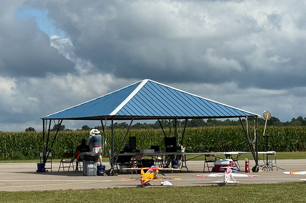 Blue canopy tent with chairs and tables on a paved area, cornfield in the background.