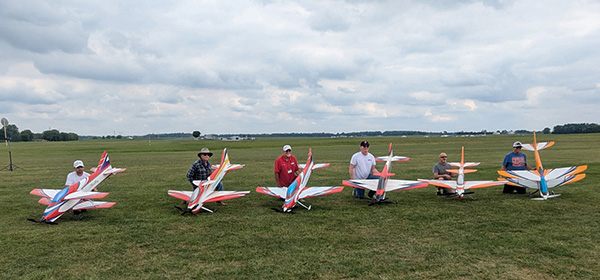 Five people with large model airplanes on a grassy field under a cloudy sky.