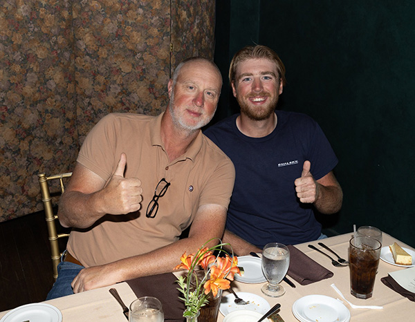 Two men seated at a table, smiling and giving thumbs up.