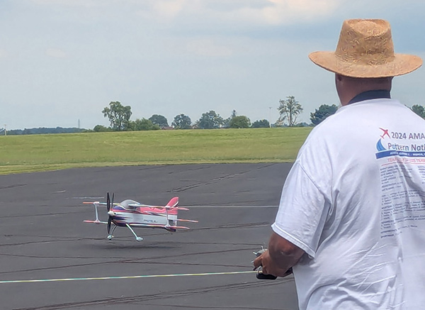 A man in a straw hat flies a model airplane on an open runway.