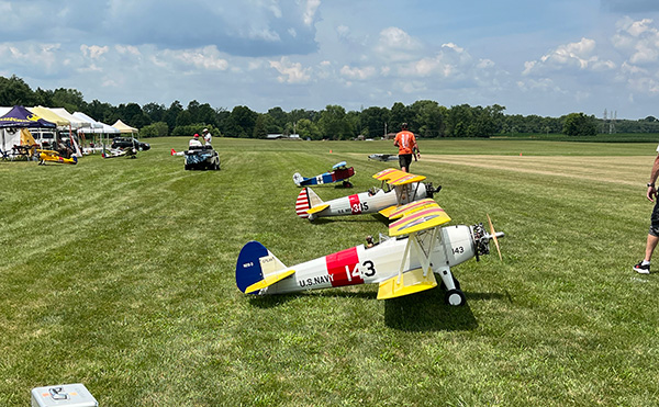 Model airplanes on grass field, tents and trees in background, clear sky.