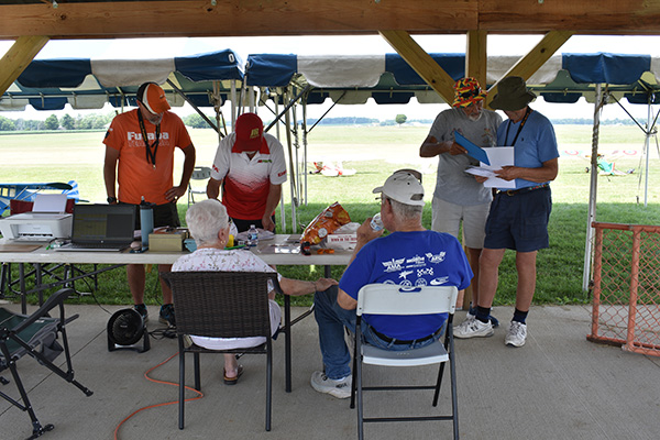 People gathered under an outdoor gazebo, some seated and reading papers.