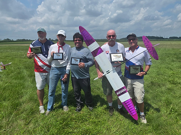 Five people holding trophies with a model glider on a grassy field.
