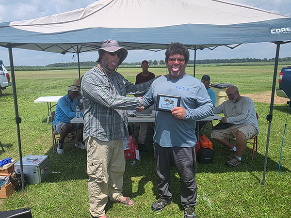 Two men shaking hands, one holding a framed photo, under a canopy at an outdoor event.