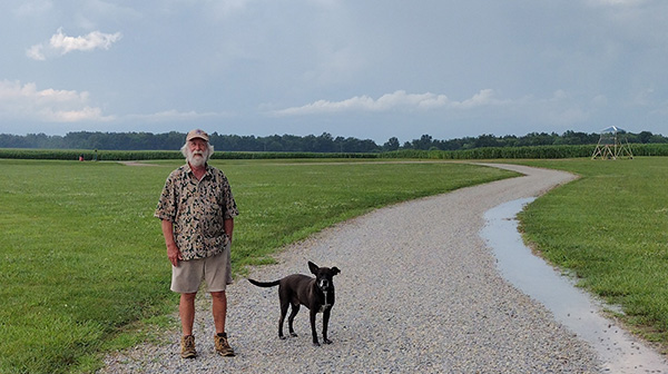 Old man with a black dog on a gravel path in a green field under a cloudy sky.