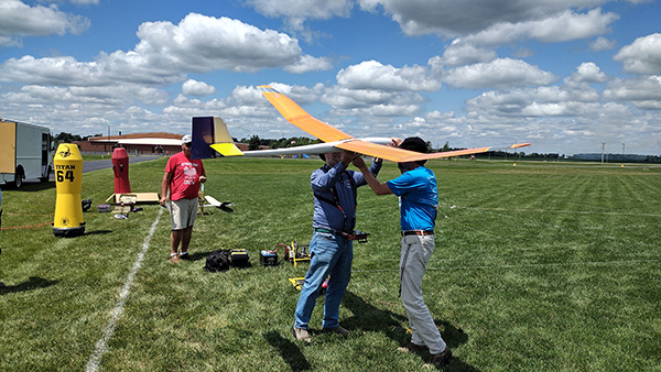 Large model glider held by two men on a grassy field, with a blue sky and clouds.