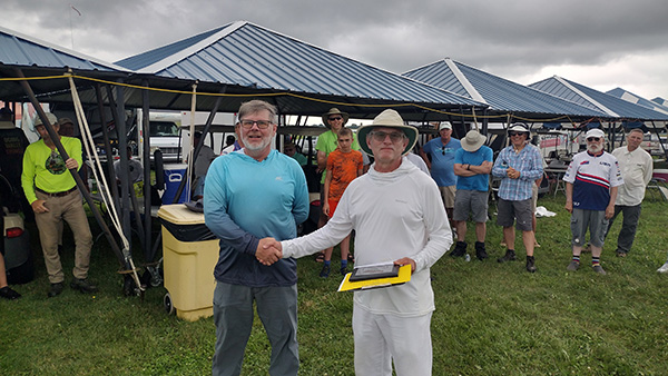 Two men shaking hands outdoors under a blue canopy. Crowd watching in the background.