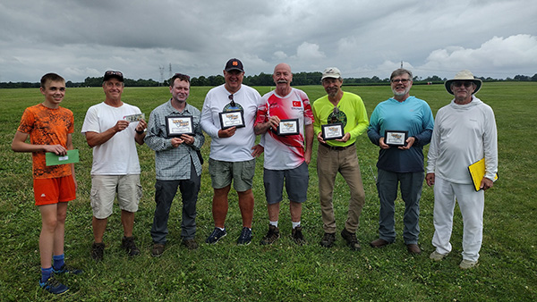 A group of eight people standing on grass, holding plaques, under a cloudy sky.