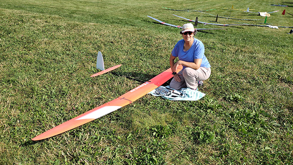 Woman squatting beside a large red model glider on a grassy field.