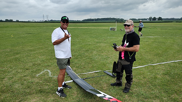 Two men in a grassy field with a model glider, one wearing sunglasses.