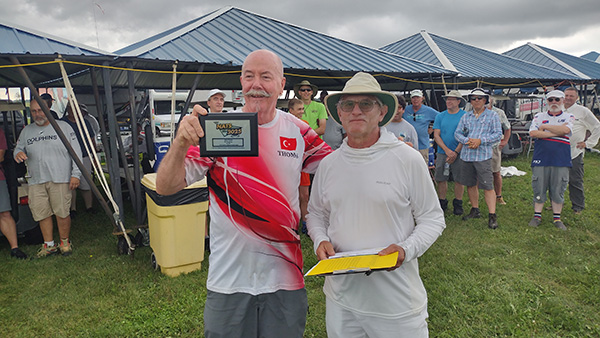 Two men holding an award, standing outdoors under a canopy.