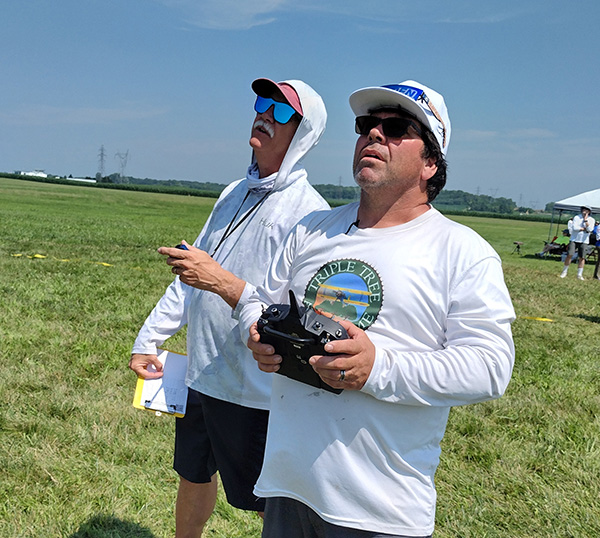 Two men operate drones on a sunny grass field.