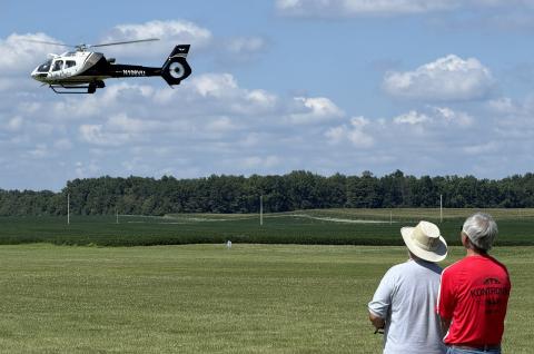 Two men watch a white helicopter flying over a grassy field.