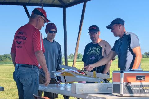 Four men examining a model aircraft on a table outdoors.