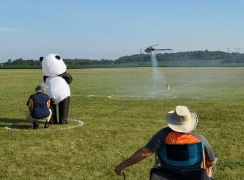 Panda mascot with two people at a field, watching a model helicopter take off.