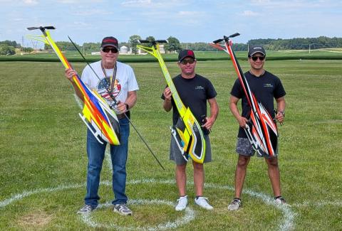 Three men smiling, holding colorful remote control helicopters on grass.