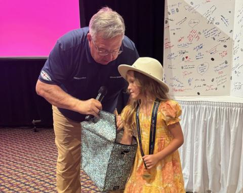 Man in blue shirt holding microphone, talking to smiling girl in yellow dress and hat.