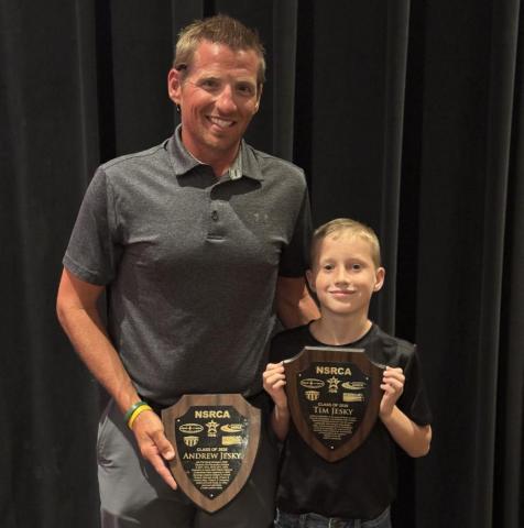Man and child smiling, holding award plaques on stage.