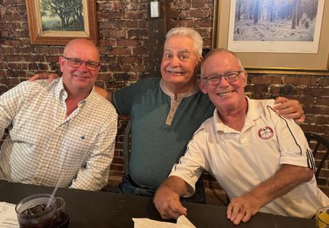 Three smiling older men sitting together at a table in a brick-walled room.