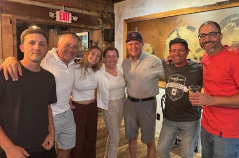 Group of seven people smiling indoors, casually dressed.